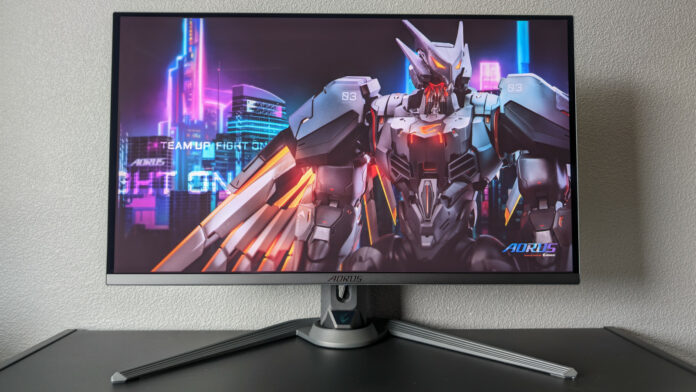 Aorus FO27Q3 gaming monitor, sat atop a black surface, against a grey background, with a bird-style mech on its screen.