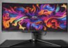 MSI MPG 341CQPX QD-OLED standing atop a black surface, against a grey background, its display brightly lit by ribbons of various colours