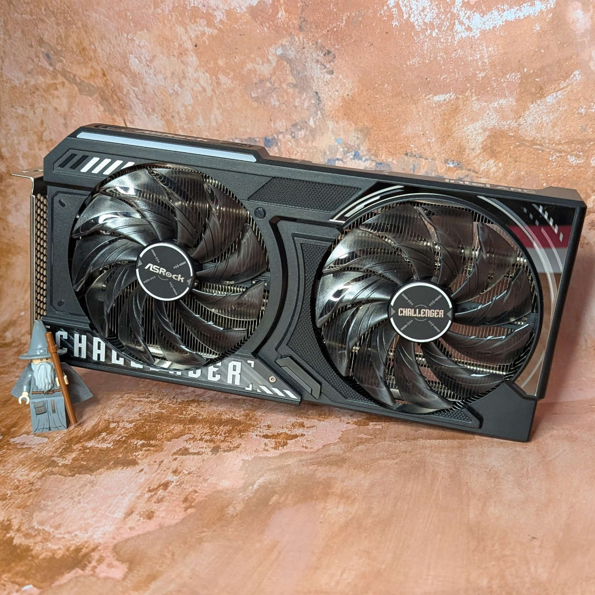 Front view of ASRock Challenger OC Arc B570 graphics card, standing upright, with a LEGO motif of Gandalf the Grey standing next to it (left).