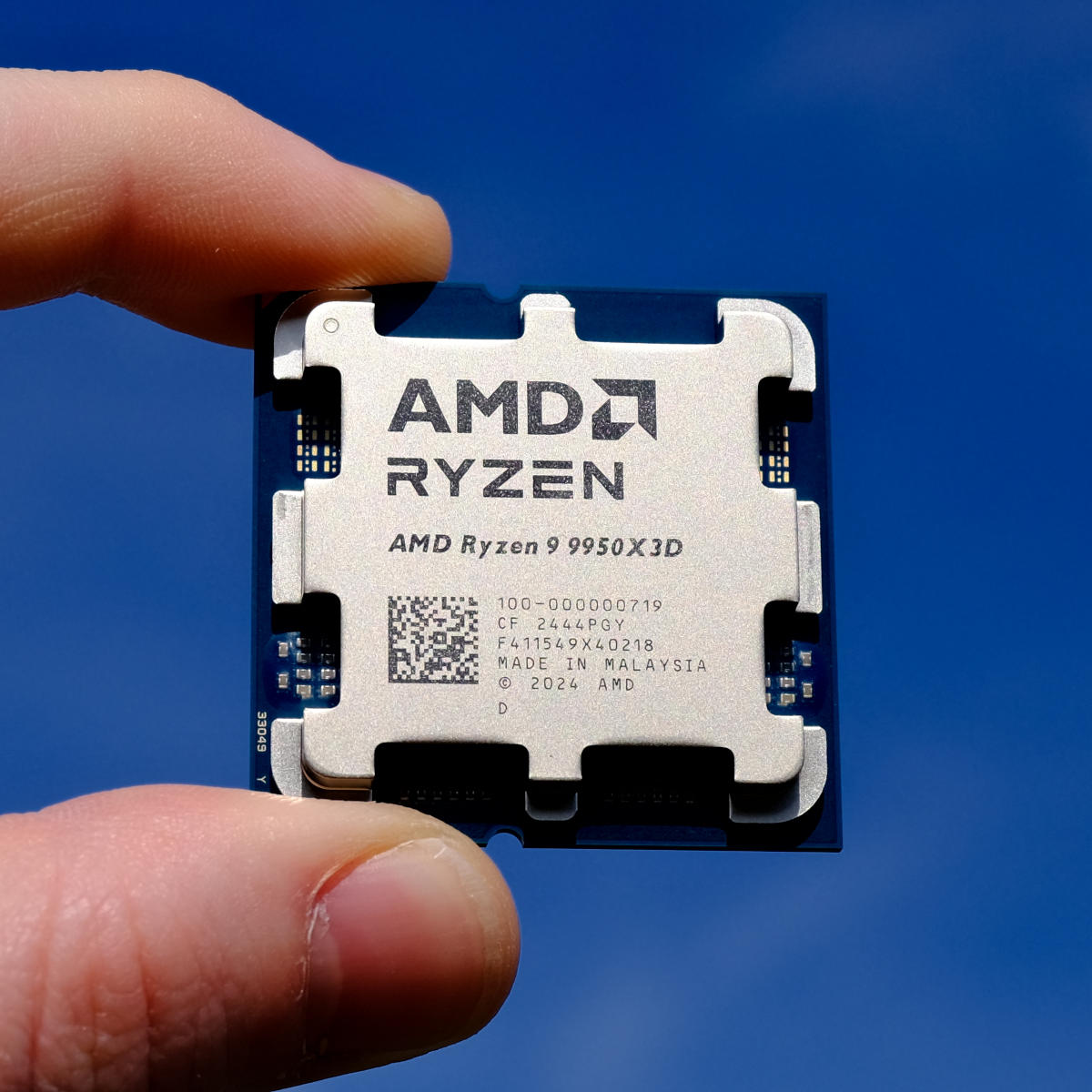 AMD Ryzen 9 9950X3D held between a finger and thumb against the backdrop of a blue sky.